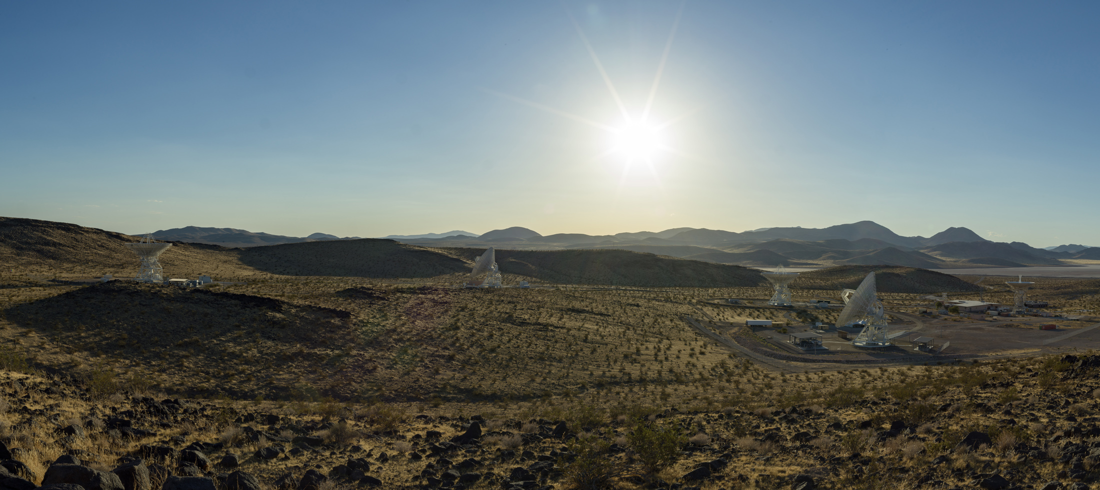 Large white Deep Space Network antennas stand in California’s Goldstone desert beneath the sun and distant mountains